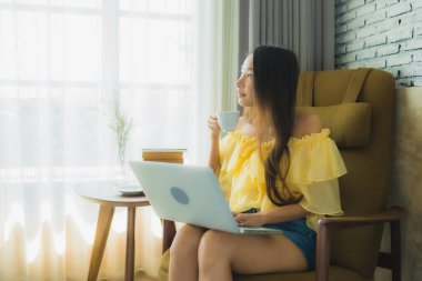 Genç Asyalı kadının portresi oturma odasında dizüstü bilgisayarı, kahve fincanı ve kitap kullanarak sandalyede oturuyor.