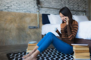 Genç Asyalı kadın yatak odasının içinde kitap ve kahve fincanı ile akıllı cep telefonu kullanıyor.