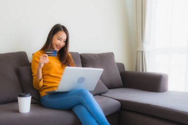 Genç Asyalı kadının oturma odasında online alışveriş yapmak için akıllı cep telefonu ve kredi kartı olan dizüstü bilgisayarı kullandığı portre.