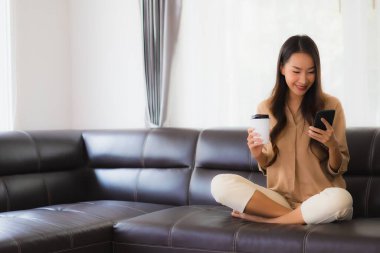Güzel bir Asyalı kadın portresi. Kanepenin iç tarafında cep telefonu ya da kahve fincanı ile cep telefonu kullanıyor.