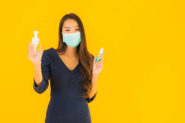 Maske, alkol jeli ve sarı izole edilmiş arka planda spreyli güzel Asyalı kadın portresi.