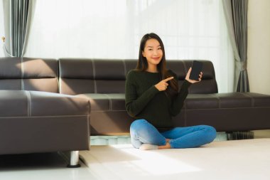 Güzel bir Asyalı kadın portresi oturma odasının iç tarafında kanepeli bir kahve fincanı ve akıllı cep telefonu kullanıyor.