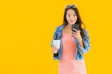 Kahve fincanı ve sarı izole edilmiş arka planda akıllı cep telefonlu güzel Asyalı kadın portresi.