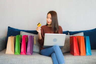 Asyalı güzel bir kadının portresi oturma odasındaki kanepede online alışveriş yapmak için bilgisayar ve kredi kartı kullanıyor.