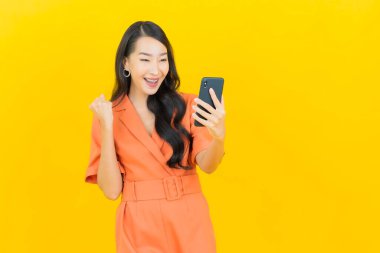 Asyalı güzel bir kadının portresi sarı arka planda akıllı cep telefonuyla gülümsüyor.