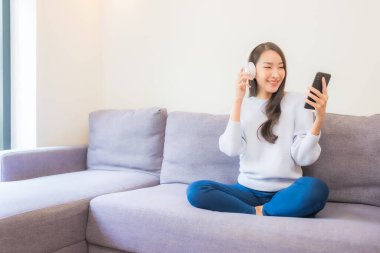 Güzel bir Asyalı kadın oturma odasında müzik dinlemek için akıllı cep telefonu kulaklığı kullanıyor.