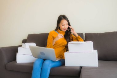 Asyalı genç bir kadının portresi, oturma odasındaki koltukta paket kutusuyla çalışmak için dizüstü bilgisayar kullanıyor.