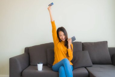 Genç Asyalı kadının oturma odasında online alışveriş yapmak için akıllı cep telefonu ve kredi kartı olan dizüstü bilgisayarı kullandığı portre.