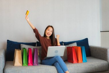 Asyalı güzel bir kadının portresi oturma odasındaki kanepede online alışveriş yapmak için bilgisayar ve kredi kartı kullanıyor.