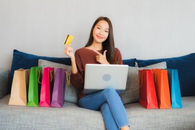 Asyalı güzel bir kadının portresi oturma odasındaki kanepede online alışveriş yapmak için bilgisayar ve kredi kartı kullanıyor.