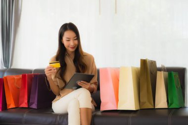 Güzel Asyalı bir kadının portresi. İnternetten alışveriş yapıyor. Kredi kartı ve akıllı telefonuyla oturma odasındaki alışveriş çantasının etrafında.