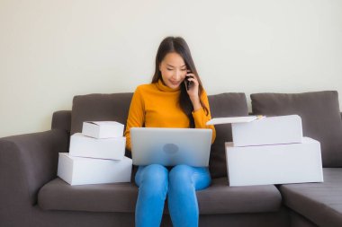Asyalı genç bir kadının portresi, oturma odasındaki koltukta paket kutusuyla çalışmak için dizüstü bilgisayar kullanıyor.