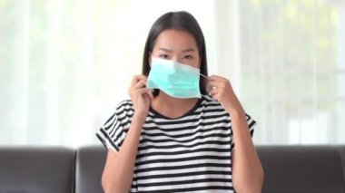 Maskeli güzel Asyalı kadın Coronavirus 'u ya da Covid19' u korumak için oturma odasında kanepede oturur.