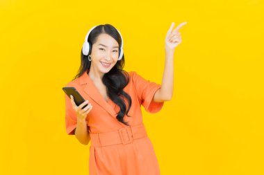 Kulaklık ve akıllı cep telefonlu güzel Asyalı kadın portresi. Sarı arka planda müzik dinliyor.