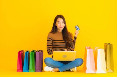 Güzel bir portre genç Asyalı kadın dizüstü bilgisayarla ya da dizüstü bilgisayarla yerde oturuyor sarı arka planda alışveriş torbası etrafında online alışveriş yapmak için kredi kartı kullanıyor.
