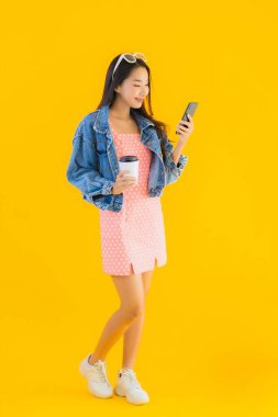 Kahve fincanı ve sarı izole edilmiş arka planda akıllı cep telefonlu güzel Asyalı kadın portresi.