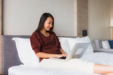 Yatak odasındaki yatakta bilgisayar ve cep telefonu kullanan güzel Asyalı kadınların portresi.