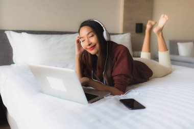 Yatak odasındaki yatakta müzik dinlemek için bilgisayar ve kulaklık kullanan güzel Asyalı kadınların portresi.