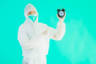 Güzel bir portre genç Asyalı doktor. Özel süit ve maske takıyor. Mavi arka planda saati ya da alarmı gösteriyor.