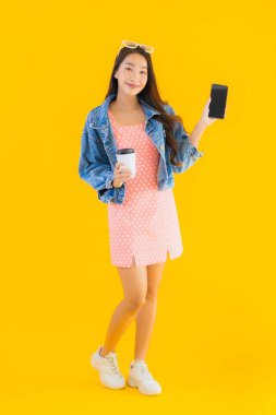Kahve fincanı ve sarı izole edilmiş arka planda akıllı cep telefonlu güzel Asyalı kadın portresi.