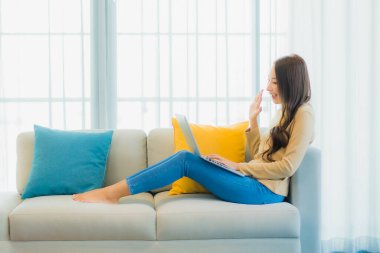 Asyalı güzel bir kadının portresi oturma odasının iç dekorasyonunda dizüstü bilgisayar kullanıyor.