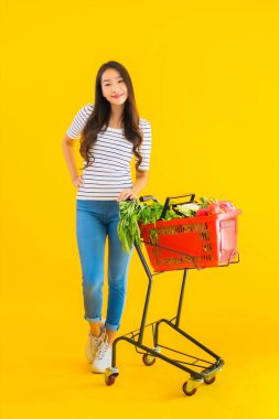 Güzel genç Asyalı kadın süpermarketten alışveriş yapıyor ve sarı izole arka planda el arabası var.