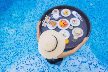 Asyalı güzel bir kadının portresi yüzme havuzunda yüzen kahvaltı tepsisiyle mutlu bir gülümseme