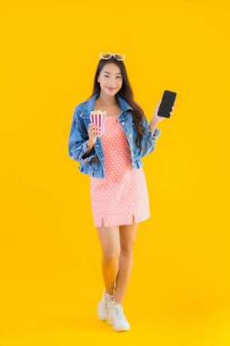 Güzel bir Asyalı kadın portresi telefon patlamış mısırı ve sarı izole edilmiş bir arka planda film izlemekten memnun.