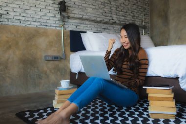 Genç Asyalı kadın yatak odasının içinde elinde kahve fincanı ve kitapla dizüstü bilgisayar kullanıyor.