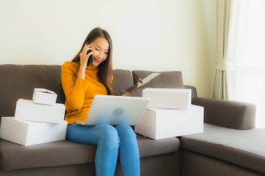 Asyalı genç bir kadının portresi, oturma odasındaki koltukta paket kutusuyla çalışmak için dizüstü bilgisayar kullanıyor.