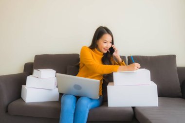Asyalı genç bir kadının portresi, oturma odasındaki koltukta paket kutusuyla çalışmak için dizüstü bilgisayar kullanıyor.