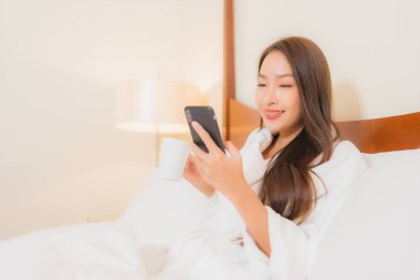 Güzel bir Asyalı kadın yatak odasının içinde akıllı cep telefonu kullanıyor.