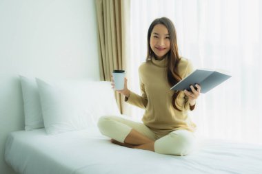 Genç Asyalı kadın, elinde kahve fincanıyla yatak odasında kitap okuyor.