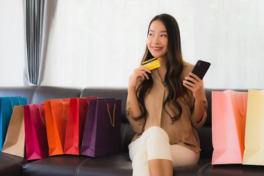 Güzel Asyalı bir kadının portresi. İnternetten alışveriş yapıyor. Kredi kartı ve akıllı telefonuyla oturma odasındaki alışveriş çantasının etrafında.