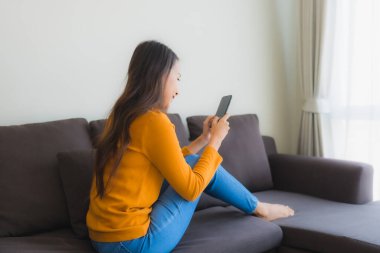 Genç Asyalı kadının oturma odasında yastıklı akıllı cep telefonu kullandığı portre.