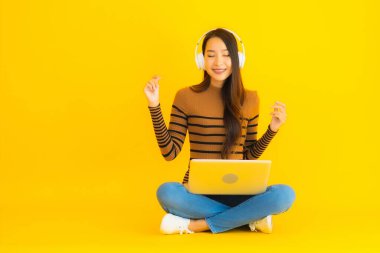 Güzel bir portre genç Asyalı kadın dizüstü bilgisayar ve kulaklıkla sarı izole arka planda oturuyor.