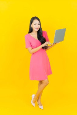 Asyalı güzel bir kadının portresi. Sarı izole edilmiş arka planda dizüstü bilgisayarla gülümsüyor.