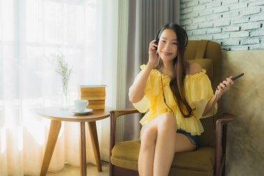 Genç Asyalı bir kadının portresi sandalyeye oturup oturma odasında cep telefonu kahvesi ve kitapla müzik dinlemek.