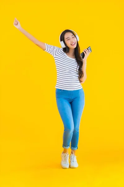 形象漂亮的年轻亚洲女人用智能手机和耳机在黄色隔离的背景下听音乐 — 图库照片