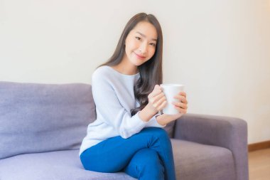 Oturma odasındaki kanepede kahve fincanı taşıyan güzel Asyalı kadın portresi.