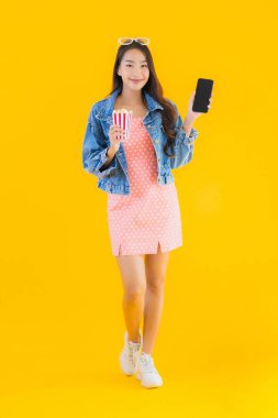 Güzel bir Asyalı kadın portresi telefon patlamış mısırı ve sarı izole edilmiş bir arka planda film izlemekten memnun.