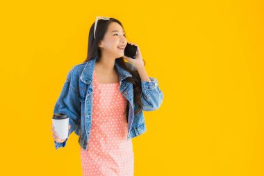 Kahve fincanı ve sarı izole edilmiş arka planda akıllı cep telefonlu güzel Asyalı kadın portresi.