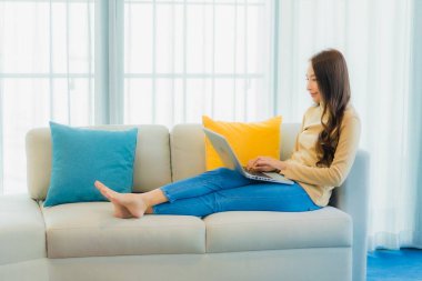 Asyalı güzel bir kadının portresi oturma odasının iç dekorasyonunda dizüstü bilgisayar kullanıyor.