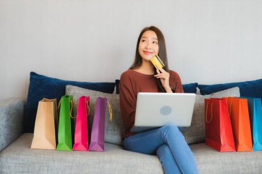 Asyalı güzel bir kadının portresi oturma odasındaki kanepede online alışveriş yapmak için bilgisayar ve kredi kartı kullanıyor.