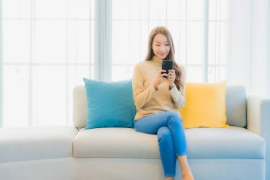 Oturma odasındaki kanepede akıllı cep telefonlu güzel Asyalı kadın portresi.