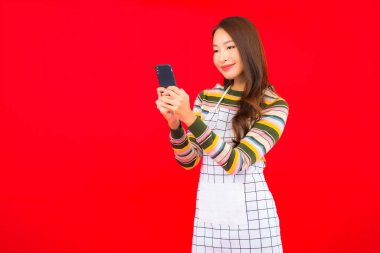Güzel bir Asyalı kadın portresi kırmızı izole edilmiş arka planda akıllı cep telefonuyla önlük giyiyor.