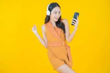 Sarı arka planda müzik dinlemek için kulaklığı ve akıllı telefonu olan güzel Asyalı kadın portresi.