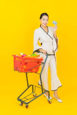 Sarı arka planda süpermarketten alışveriş sepeti almış güzel genç Asyalı kadın portresi.