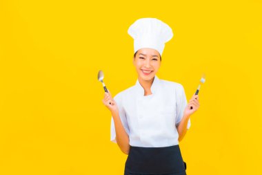 Güzel bir portre genç Asyalı kadın aşçı ya da aşçı üniforması giyiyor sarı izole arka planda çatal ve kaşıkla.