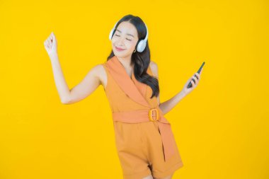 Sarı arka planda müzik dinlemek için kulaklığı ve akıllı telefonu olan güzel Asyalı kadın portresi.